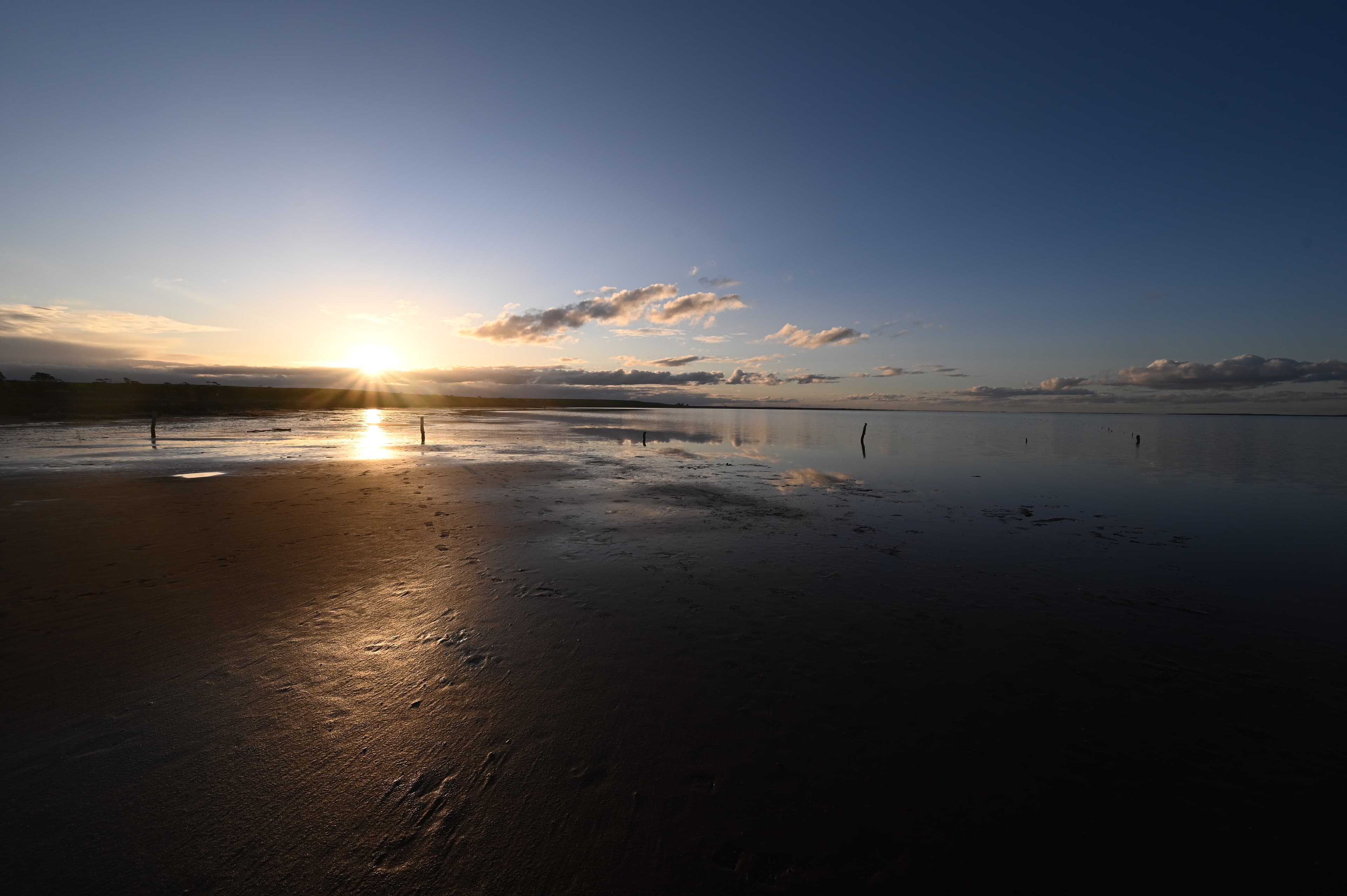 The sun sets over the clouds and the mirror-like surface of Lake Tyrrell, casting warm hues across the landscape. The reflections blur the line between sky and water.