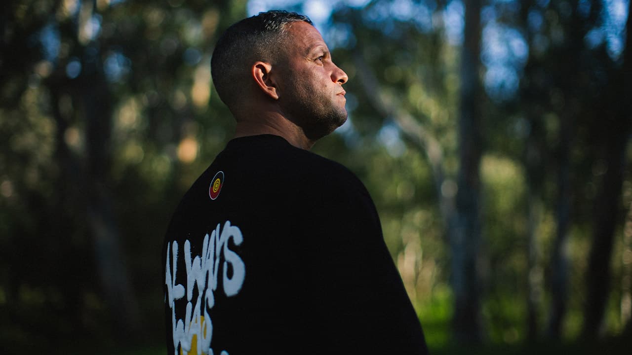 Jarvis Atkinson stands facing a view of softly out-of-focus blurred lush green trees, wearing a t-shirt with the words “Always Was” printed on the back