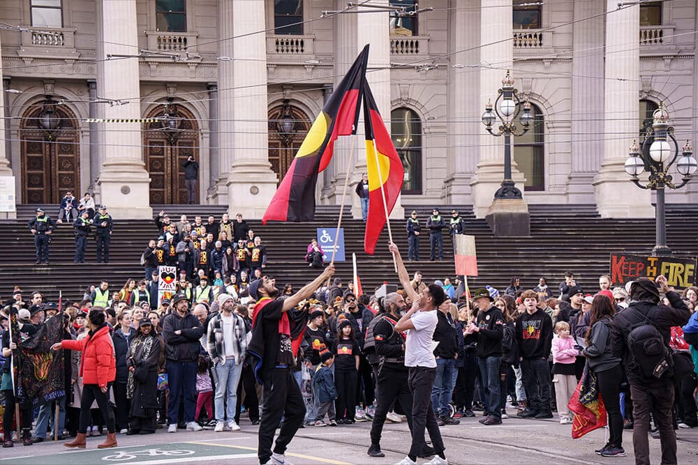 A crowd watches and cheers in front of Parliament House as two people point aboriginal flags to the sky.