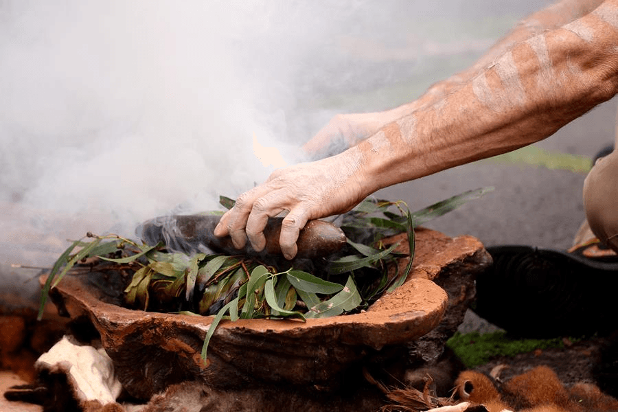 Gum leaves billow smoke from a wooden bowl. Two arms adorned with traditional body art, reach in and tend to the smoke, during a Welcome to Country smoking ceremony.