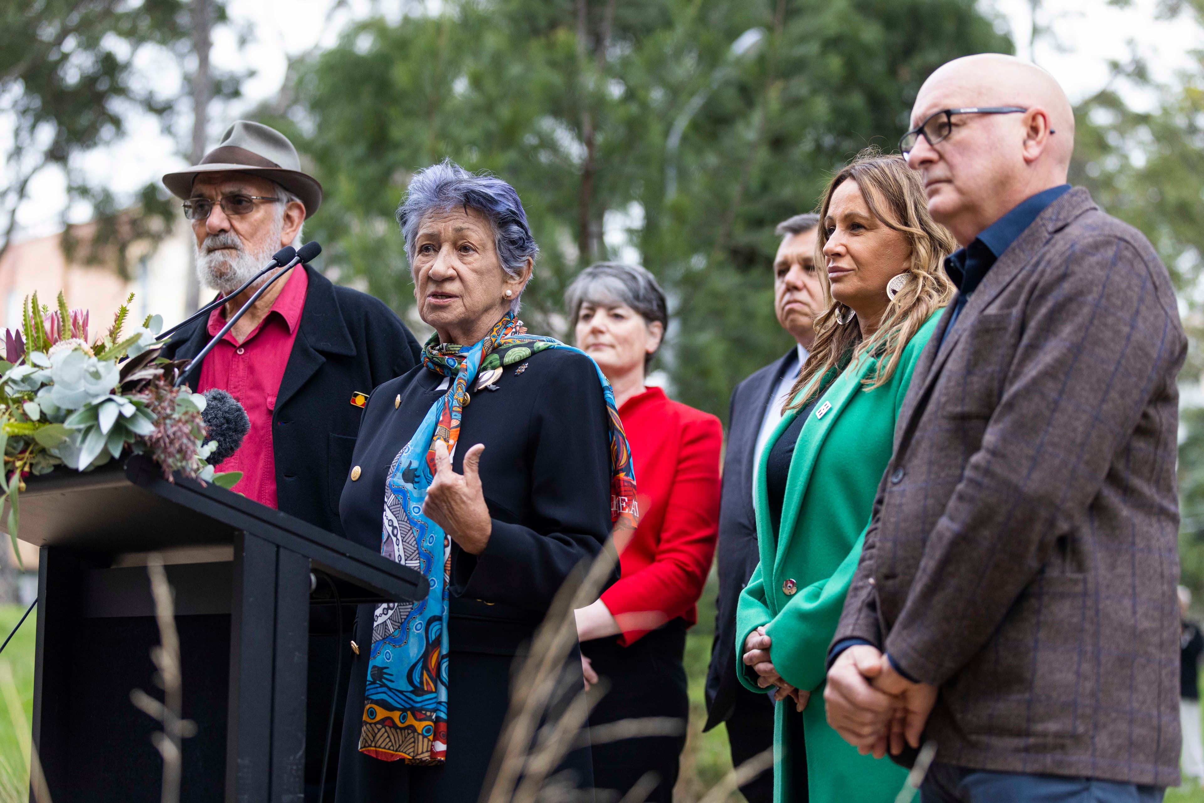 Respected Wergaia/Wamba Wamba Elder Aunty Eleanor Burke stands outdoors at a podium, delivering a speech at the opening Yoorrook Ceremony. Behind her, fellow Yoorrook commissioners and staff stand attentively, framing the moment with solidarity and shared purpose in the truth-telling journey.