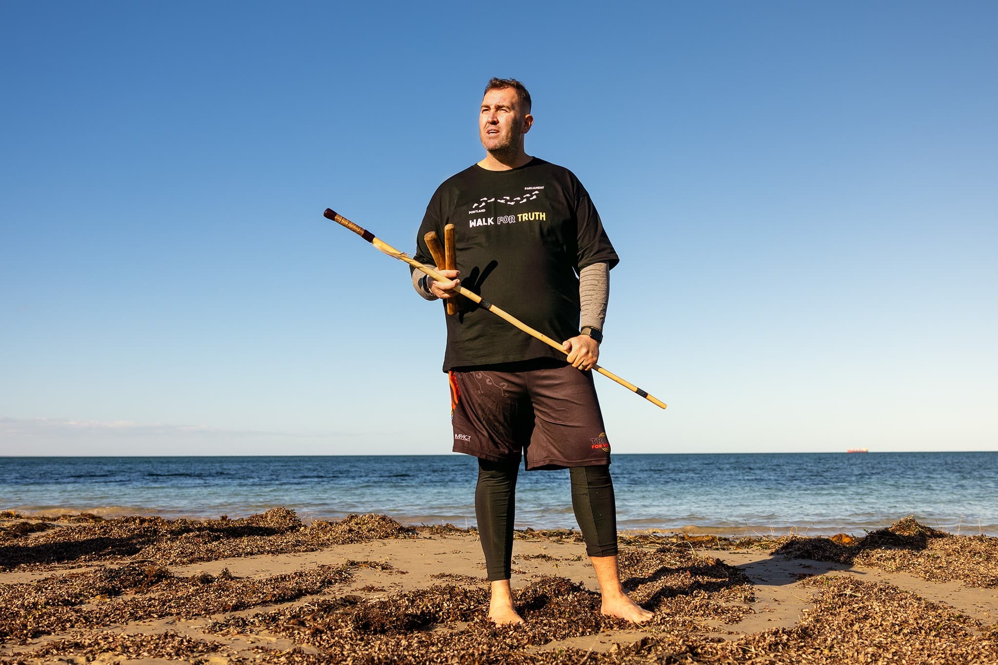 Kerrupmara/Gunditjmara man and Yoorrook Commissioner Travis Lovett, wearing a Walk for Truth t-shirt, stands on a beach with the ocean behind him at the convincing ground massacre site.