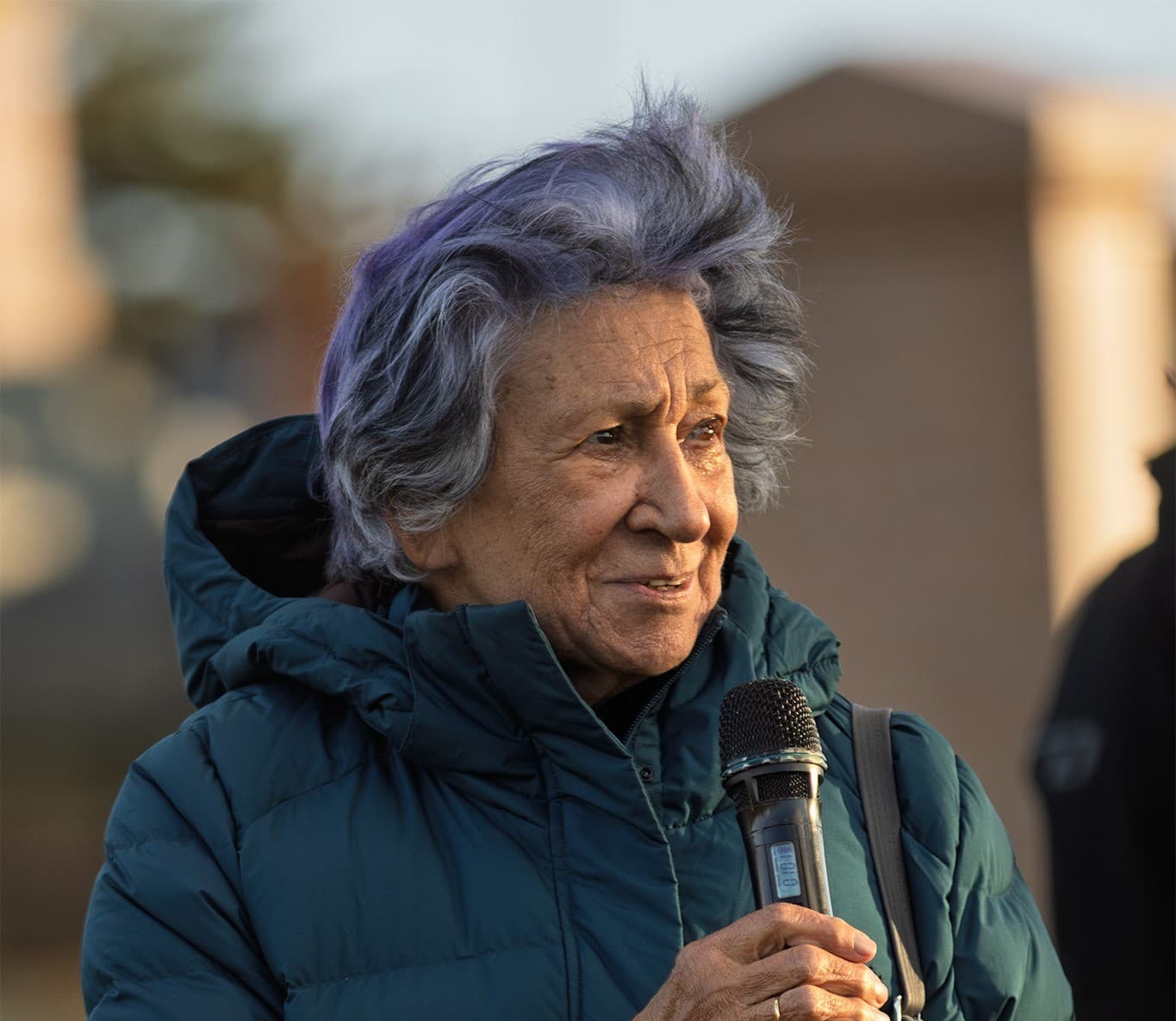 Aunty Eleanor Bourke speaks into a microphone outdoors; her purple hair is blown in the wind and the background is blurred.