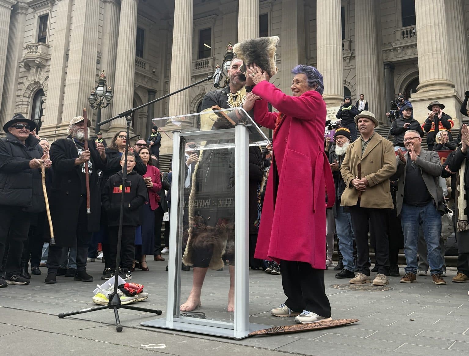 Aunty Eleanour Bourke and Travis Lovett hold up a possum skin cloak at a lectern while surrounded by a crowd on the steps of parliament house.