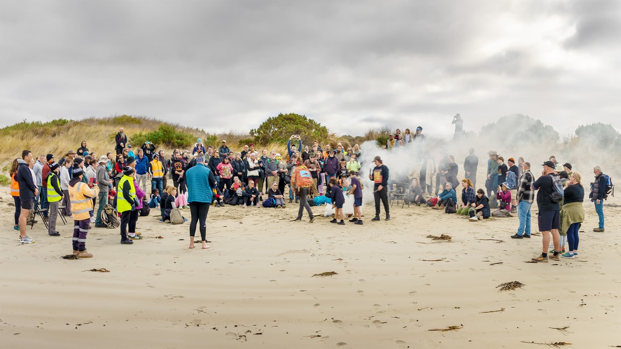 A group of people surround a smoking ceremony at a Welcome to Country been performed on a beach near Portland.