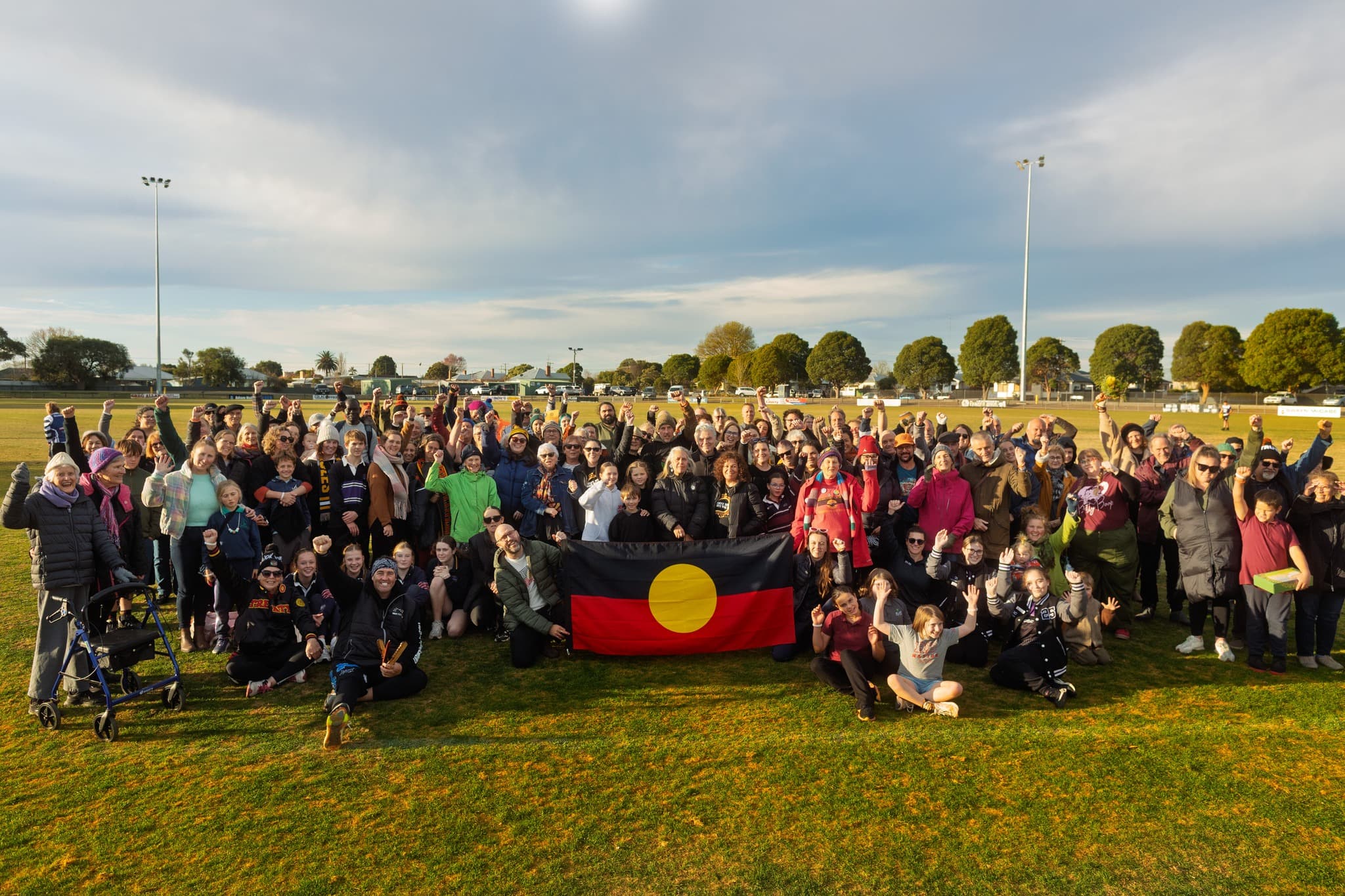 A large crowd of people stand together in solidarity around an aboriginal flag on a grassy oval.
