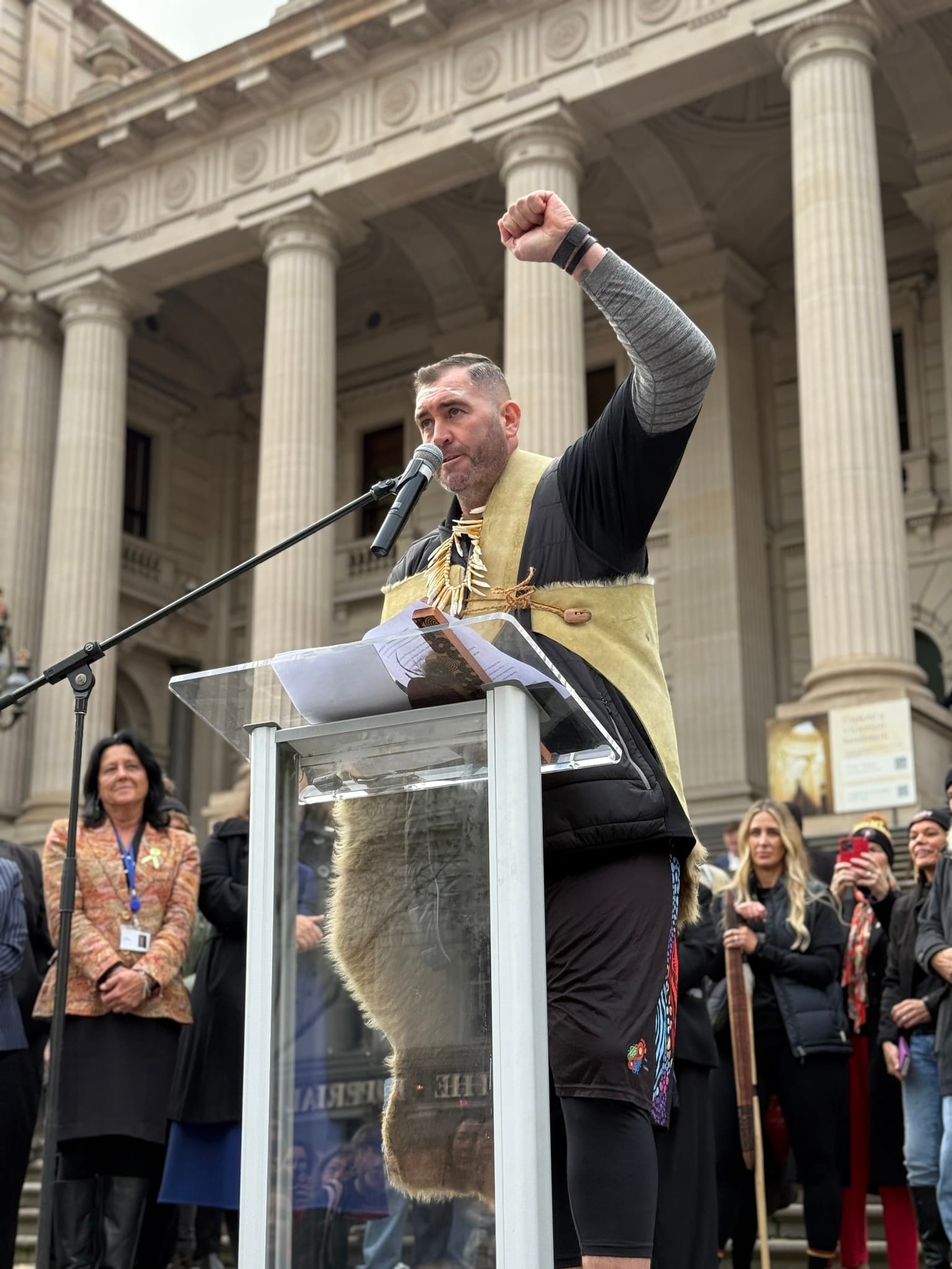 Kerrupmara/Gunditjmara man and Yoorrook Commissioner Travis Lovett, stands in front of a lectern in a possum skin cloak on the steps of parliament house. His left hand is raised high in a fist and message sticks can be seen on the lectern infront of him.