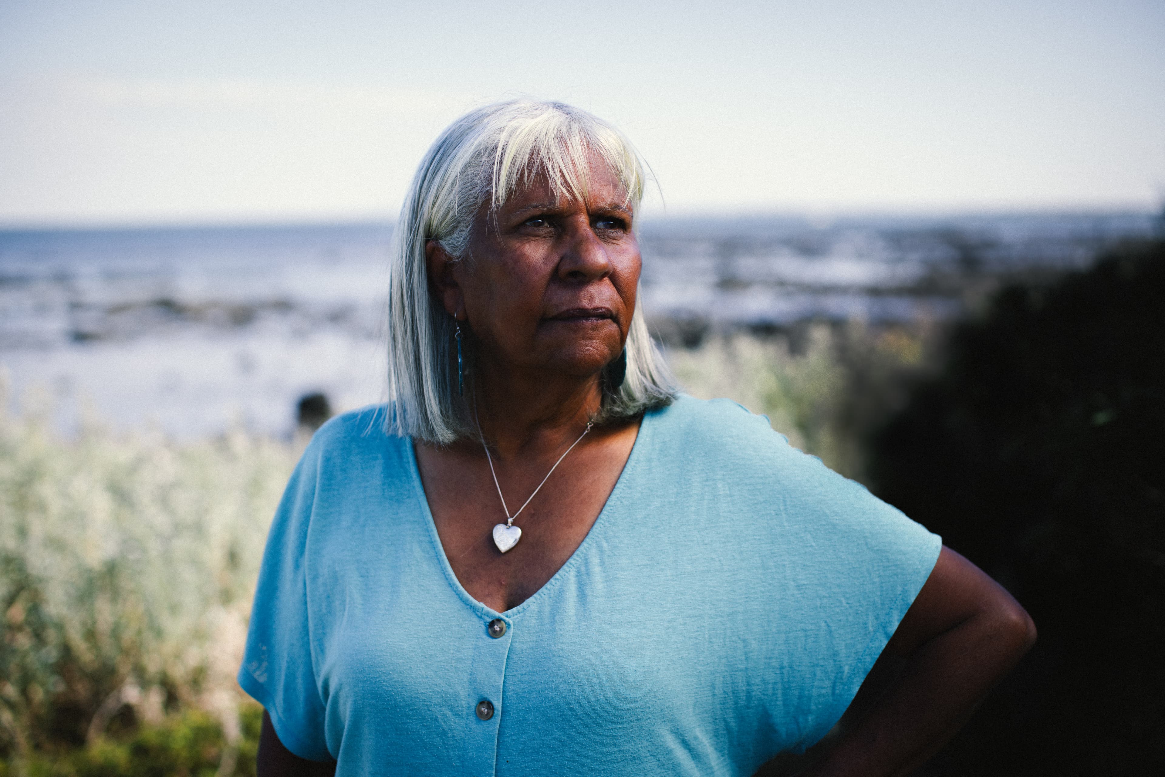 Headshot of Boon Wurrung/Bunurong, Yorta Yorta, and Mutti Mutti woman Aunty Eva-Jo Edwards, gazing thoughtfully to the left. Behind her, the ocean and coastal shrubs are slightly out of focus.