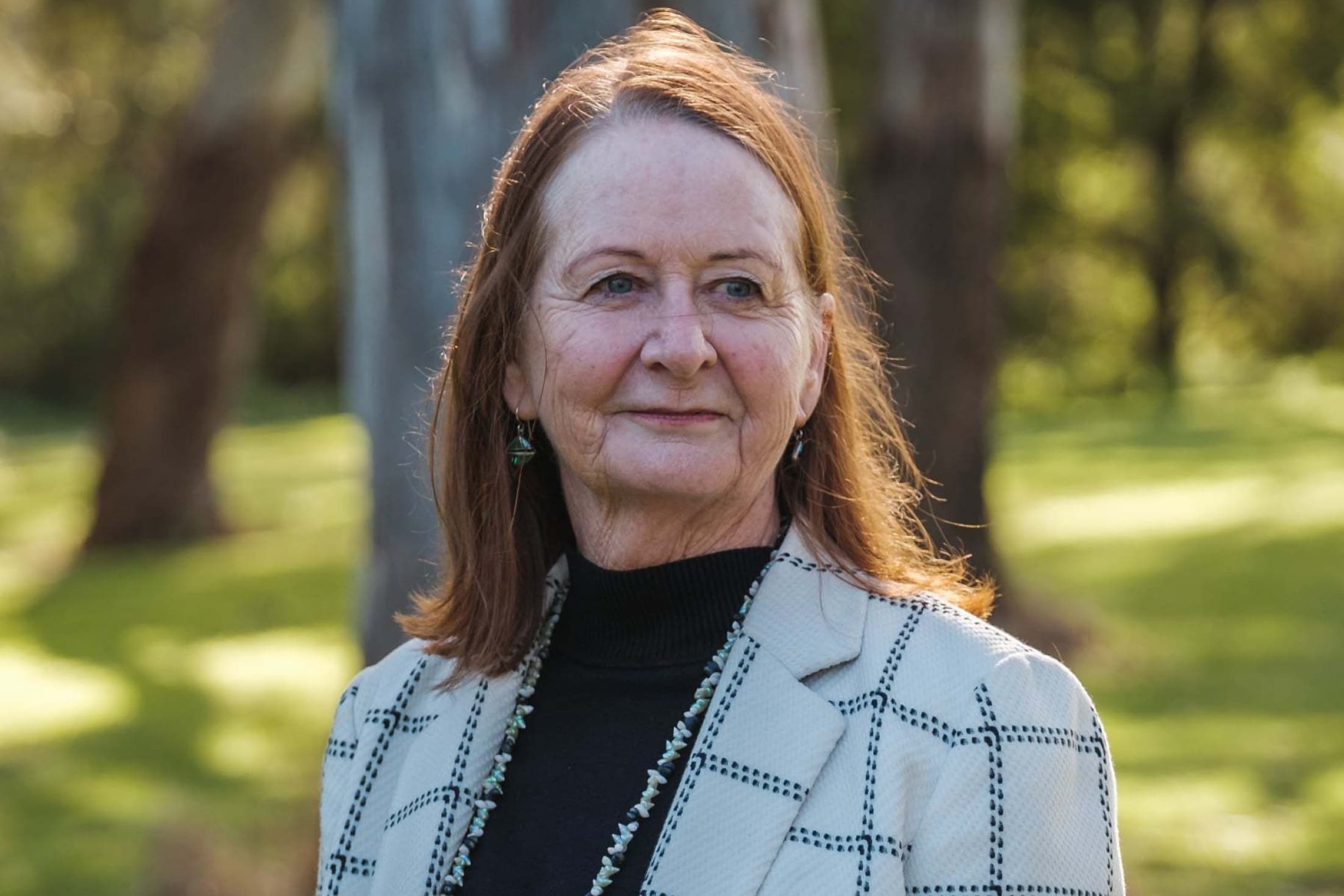 Headshot of Distinguished Professor Maggie Walter, silhouetted by large gum trees in the bacgrkound.