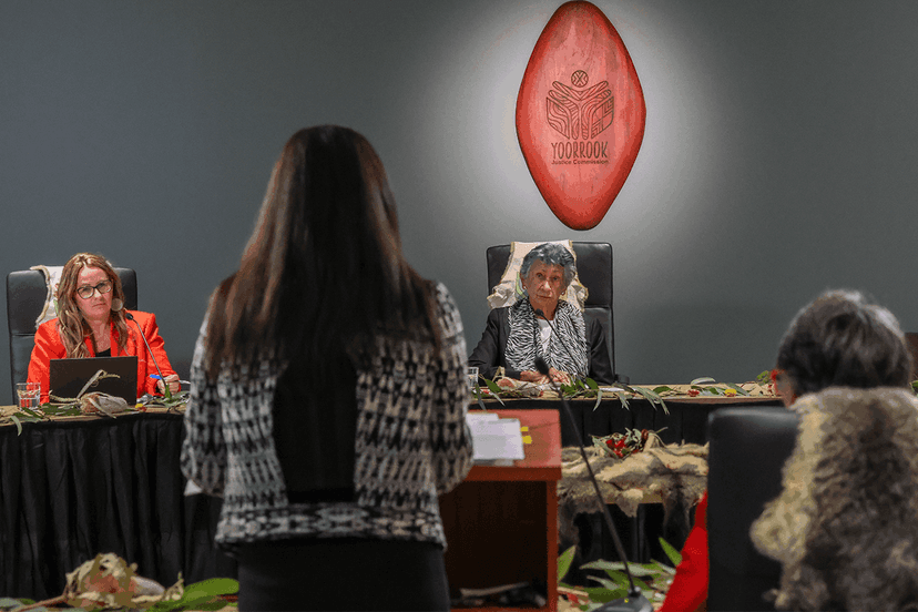 Aunty Eleanor Bourke and Sue-Ann Hunter sit at a table in front of the Yoorrook shield. They are questioning a state lawyer standing at a lectern in front of them at a directions hearing.