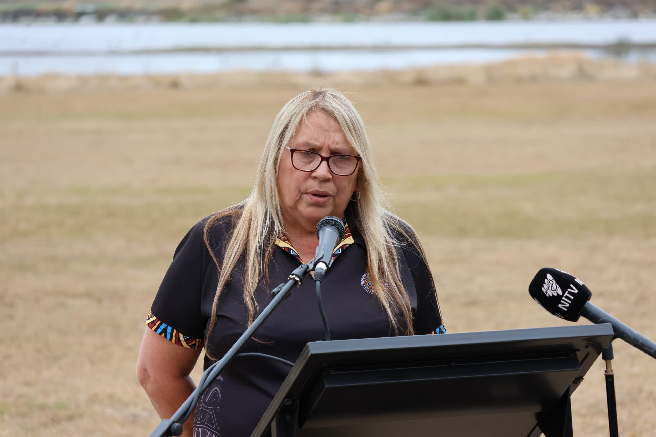 Aunty Donna Wright speaks into a microphone outdoors at a lectern. Lake Condah and a brown grassy field can be seen behind her.