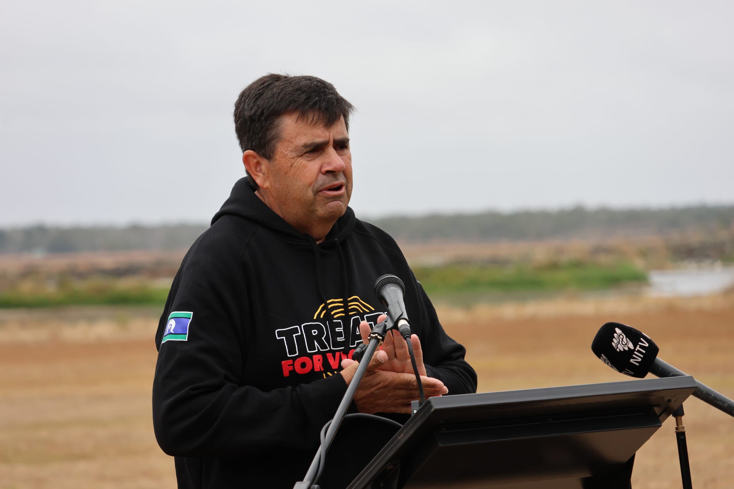 Uncle Michael (Mookeye) Bell, wearing a 'Treaty for Victoria' jumper, stands outdoors behind a lectern. Lake Condah and brown grassy fields can be seen slightly blurred behind him.