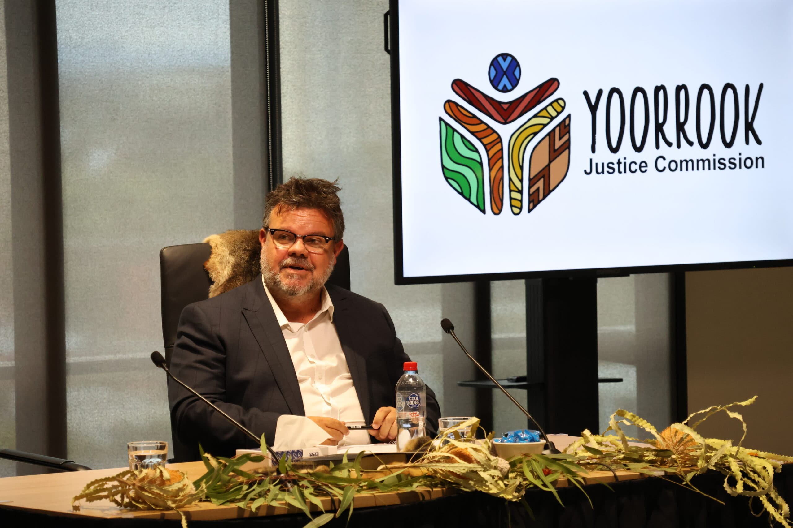 Damian Griffis from First Peoples Disability Network Australia, seated in a black chair, behind a table with microphones and adorned with bottle brush leaves. Behind him is a screen displaying the Yoorrook logo.