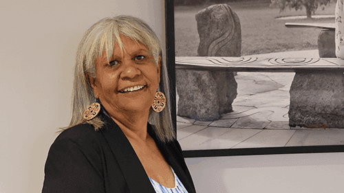 Aunty Eva Joy smiles and stands next to a photo of an Aboriginal gathering place.
