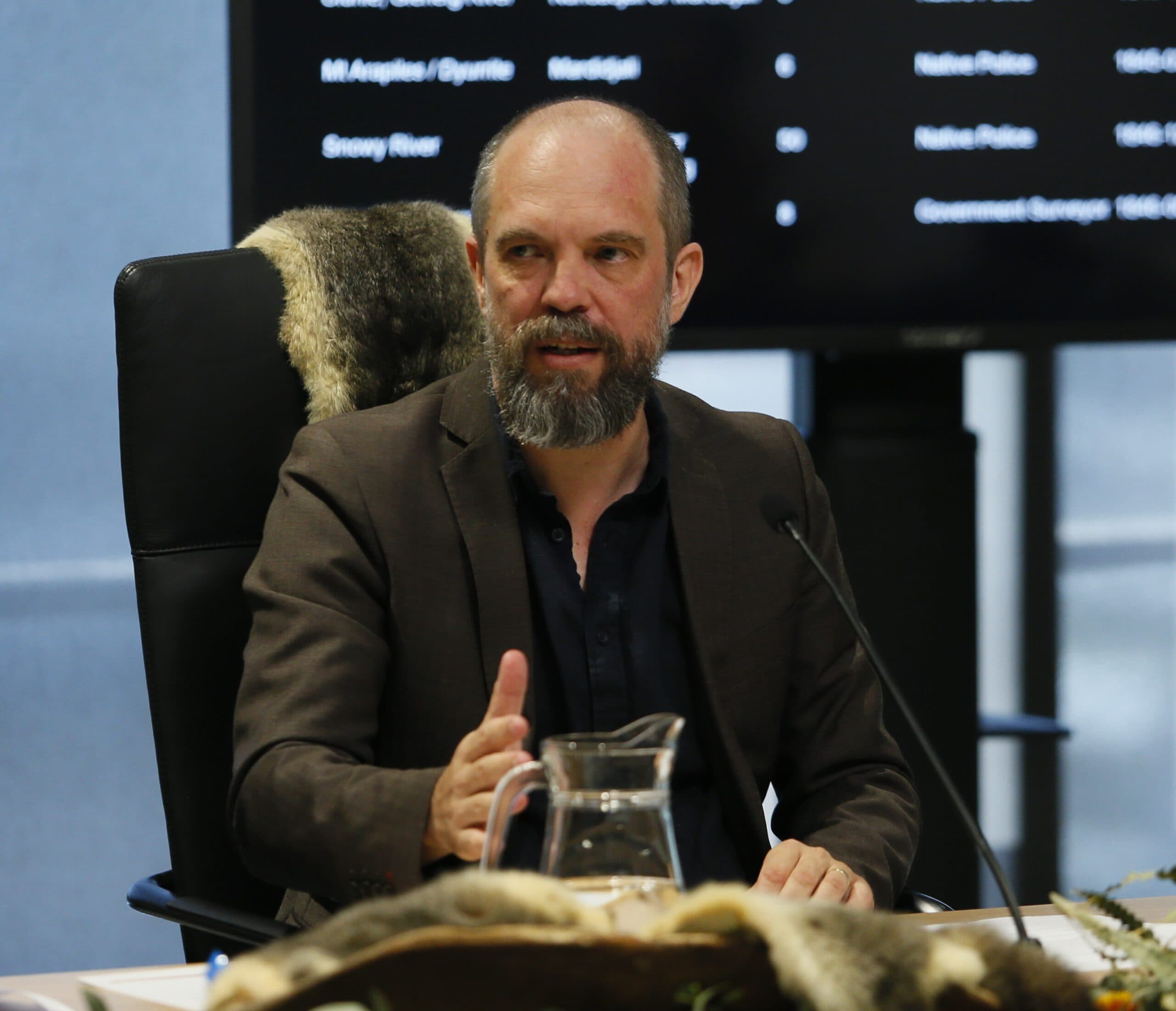 Dr Bill Pascoe from the University of Newcastle, sits at a black chair and gestures towards a microphone. A display with reported massacres can be seen behind him.