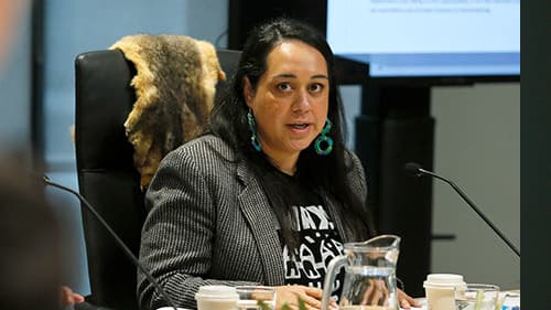 Hayley McQuire from the National Indigenous Youth Education Coalition is seated at a table with cups and a glass pitcher, indoors at a Yoorrook public hearing
