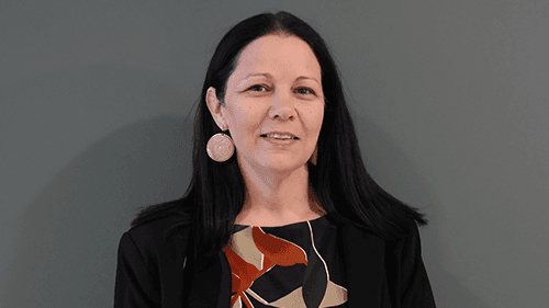 Karinda Taylor, CEO, First Peoples' Health and Wellbeing, smiles and stands in front of a grey wall.