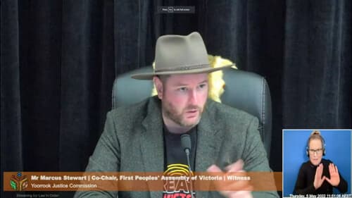 Marcus Stewart, Co-Chair, First Peoples' Assembly of Victoria, wearing a First Peoples Assembly t-shirt and Akubra hat, indoors at a Yoorrook public hearing.