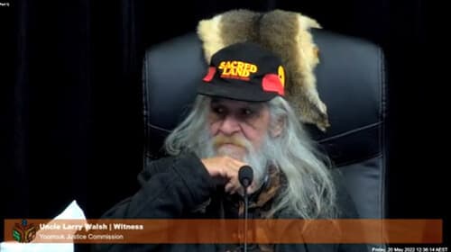 Uncle Larry Walsh, seated indoors on a black chair and wearing a cap that reads 'Sacred Land', during the Yoorrook Elders' hearings.