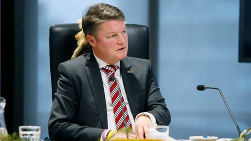 The Hon. Ben Carroll MP, Deputy Premier and Minister for Education wearing a grey suit with red and silver tie sits indoors at a chair in a Yoorrook public hearing.