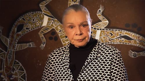 Dr Lois Peeler, wearing a black and white patterned jacket over a turtleneck, and standing in front of a wall with a traditional Indigenous painting of yellow and white lizards on a brown background