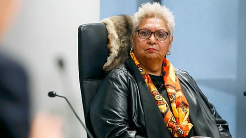 Aunty Nellie Flagg, wearing a leather jacket, and colourful yellow and orange scarf, looks off to her right while seated in a black chair at a Yoorrook public hearing.