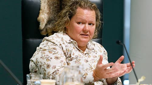 Sheree Lowe from VACCHO, seated indoors on a black chair, gestures towards a microphone at a Yoorrook public hearing