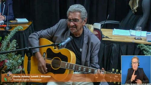 Uncle Johnny Lovett plays a guitar indoors at a Yoorrook public hearing. A sign language interpreter is overlayed in the bottom right.