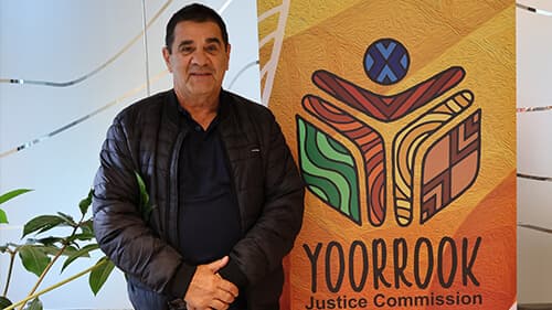 Uncle Ross Morgan stands indoors between a plant and a banner featuring the Yoorrook logo.